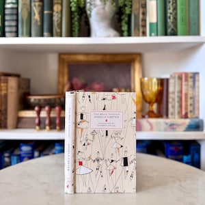 May include: A copy of "The Magic Toyshop" by Angela Carter, featuring a cream-coloured cover with a repeating pattern of figures. The spine displays the title and author's name. The book is standing upright on a table. The book is surrounded by other books.