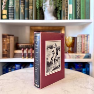 Puede incluir: Un libro de tapa dura titulado "The History of Tom Jones, A Foundling" de Henry Fielding. El libro tiene una cubierta burdeos con una ilustración enmarcada de un hombre. El lomo es azul oscuro con letras doradas.