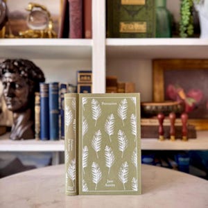 Puede incluir: Un libro de tapa dura titulado "Persuasión" de Jane Austen. El libro tiene una cubierta verde salvia con un patrón de plumas blancas. El lomo es visible y el libro está de pie sobre una superficie blanca.