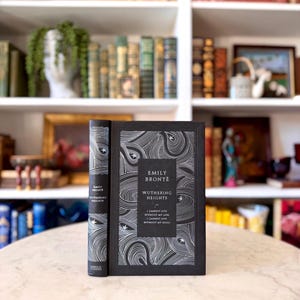 Puede incluir: Un libro de tapa dura titulado "Wuthering Heights" de Emily Brontë. El libro tiene una cubierta negra con patrones de remolinos blancos y el título en un recuadro. La cita "I cannot live without my life! I cannot live without my soul!" está impresa en la portada.