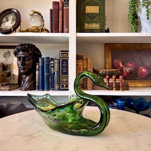 May include: A green glass swan-shaped bowl with a long, curved neck and a flared base. The bowl is a vibrant shade of green with hints of yellow and sits on a white marble surface. The swan's head is detailed, and the bowl is likely decorative.