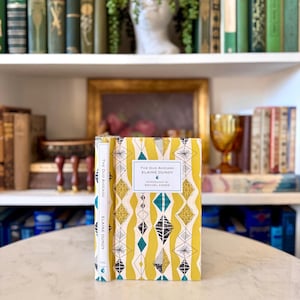 Puede incluir: Un libro de tapa dura titulado "The Dud Avocado" de Elaine Dundy. El libro tiene una cubierta amarilla con un patrón geométrico en verde azulado, negro y blanco. El lomo es blanco con el título y el nombre del autor. El libro se exhibe sobre una mesa con otros libros al fondo.