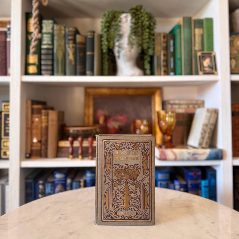 May include: A vintage book titled "Prince of the House of David" with gold and brown embossed cover art. The book is standing on a marble table in front of a bookshelf filled with antique books and decorative items.