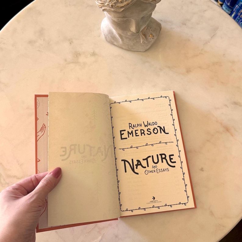 May include: An open book titled "Nature and Other Essays" by Ralph Waldo Emerson. The book has a red border and the title is in a decorative font. The book is on a marble surface with a bust in the background.