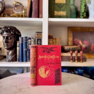 Puede incluir: Un libro antiguo titulado "About Old Storytellers" con una cubierta roja y letras doradas. El libro presenta una ilustración de un águila y una persona leyendo. El lomo es de color marrón claro con letras doradas. El libro se exhibe sobre una superficie blanca.