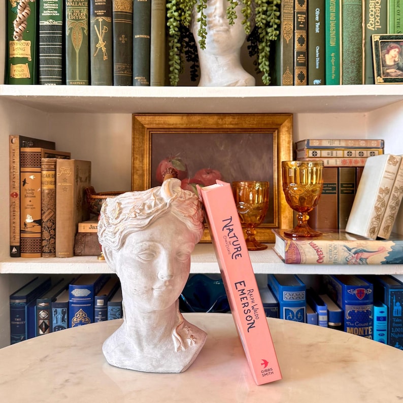 May include: A white bust of a woman with gold accents sits on a marble table, next to a pink book titled "Nature" by Ralph Waldo Emerson. The background features a bookshelf filled with vintage books and decorative items.