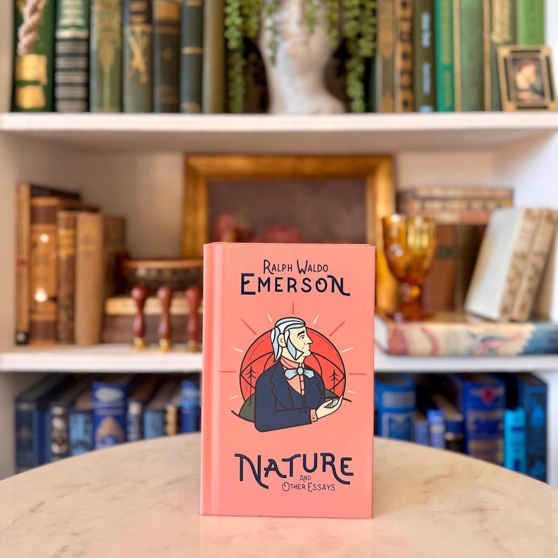 May include: A coral-colored book titled "Nature and Other Essays" by Ralph Waldo Emerson. The cover features an illustration of a man in a blue coat and a white cravat, set against a backdrop of trees and a sunburst design. The book is on a table.