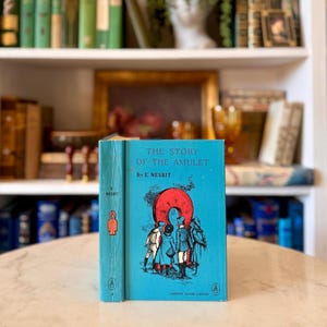 May include: A vintage hardcover book titled "The Story of the Amulet" by E. Nesbit. The book has a turquoise cover with an illustration of people and a red circle. The spine is turquoise with the author's name and a small illustration.