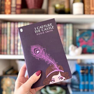 Peut inclure: Un livre de poche intitulé "I Capture the Castle" de Dodie Smith. La couverture est violet foncé avec une plume de paon rose et un visage de femme. Le livre est tenu dans une main, avec des étagères en arrière-plan.