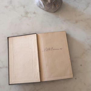 May include: An open book with aged, off-white pages. The left page is blank, while the right page has handwritten cursive text that reads "R.B. Annie". The book has a dark grey cover and spine.