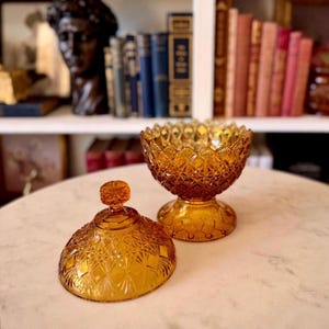 May include: An amber-colored glass candy dish with a matching lid, featuring a pressed diamond pattern and a scalloped rim. The bowl sits on a pedestal base, and the lid has a small, round knob handle, showcasing a vintage-style pressed glass design.