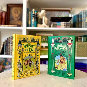 Könnte beinhalten: Zwei Hardcover-Bücher aus der Wizard of Oz-Reihe. Das gelbe Buch trägt den Titel "The Wizard of Oz: The First Five Novels". Das grüne Buch trägt den Titel "The Emerald City of Oz: Novels Six Through Ten of the Oz Series". Beide Bücher haben Illustrationen.