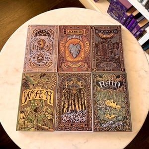 May include: Six hardcover books from the Blackwater series by Michael McDowell. The covers feature intricate illustrations in shades of brown, gold, and green. Titles include "Blackwater I", "Blackwater II", "Blackwater III", "Blackwater IV", "The Fortune", and "Rain".