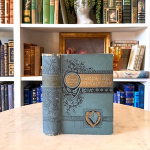 May include: Antique book titled "Harrison and Morton" with a teal cover and gold lettering. The book has decorative floral and shield designs. The spine reads "Harrison & Morton" in embossed lettering. The book is displayed on a marble surface.