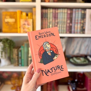 May include: A coral-colored book titled "Nature and Other Essays" by Ralph Waldo Emerson. The cover features an illustration of Emerson in a blue coat and white hair, set against a red and orange background. The book is held in front of a bookshelf.