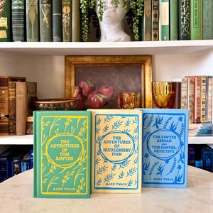 May include: Three vintage hardcover books by Mark Twain: "The Adventures of Tom Sawyer," "The Adventures of Huckleberry Finn," and "Tom Sawyer Abroad and Tom Sawyer, Detective." The books are in green, cream, and blue with gold and blue lettering.