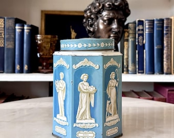 May include: A hexagonal light blue metal tin featuring raised white relief figures of classical deities in arched panels. The lid has a decorative floral border. Each panel includes a name plate, with the center figure labeled as RHEA.