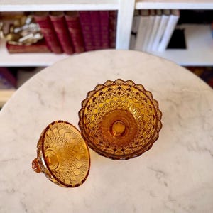 May include: An amber-colored glass candy dish with a matching lid, featuring a pressed diamond and starburst pattern. The bowl has a scalloped rim, and the lid includes a small knob handle, both showcasing intricate geometric glass textures.