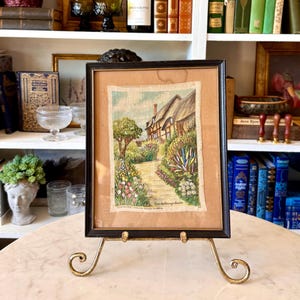 May include: Framed artwork depicting a cottage scene with a path leading to the house, surrounded by colorful flowers and greenery. The artwork is in a black frame and displayed on a gold-colored easel.
