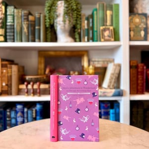 May include: A hardcover book titled "Alice's Adventures in Wonderland and Through the Looking Glass" by Lewis Carroll. The book has a pink cover with whimsical illustrations and a matching pink spine. The book is displayed on a round table.