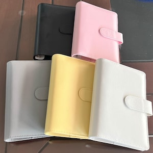 May include: Five different colored leather-bound journals with a flap closure and a metal clasp. The journals are in black, pink, gray, yellow, and white.