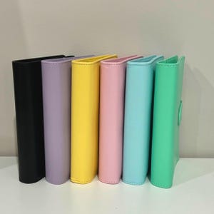 May include: A row of six colorful binders in black, lavender, yellow, pink, light blue, and green. The binders are standing upright on a white surface. Each binder has a smooth, solid color and a rounded spine.