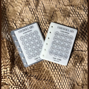 May include: Two clear plastic sheets with a savings challenge tracker. The top sheet has a goal of £2,000 and the bottom sheet has a goal of £2. Each sheet has a grid of circles with numbers printed inside. The top sheet has 100s and the bottom sheet has 10s.
