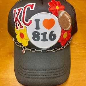 May include: Black trucker hat with a variety of patches, including "KC", "I love 816", a red flower, and a football. The hat has a silver chain with a charm.
