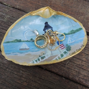 May include: A seashell dish painted with a lighthouse and a sailboat on a blue ocean. The dish is decorated with gold paint and has the words "Cape Cod, MA" written on the side. The dish is filled with jewelry, including two rings and a necklace.