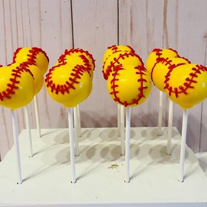 May include: A set of yellow and red softball cake pops on white sticks. The cake pops are arranged on a white display stand.
