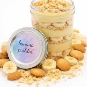 May include: A glass jar filled with banana pudding, layered with sliced bananas, vanilla wafers, and crumbled cookies. A small, round container with a lid that reads "banana puddin'" sits next to the jar. Scattered around are more cookies and banana slices.