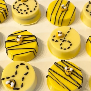 May include: Close-up of several yellow and white frosted treats decorated with black stripes and small bee-shaped candies. The treats are round and appear to be cookies or candies, perfect for a themed party or gift.