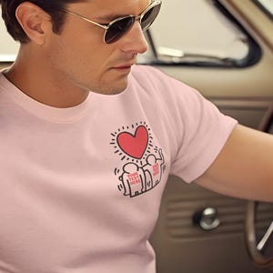 May include: A man wearing a pink t-shirt with a red heart and a graphic of people holding hands. He is wearing sunglasses and sitting in a car.