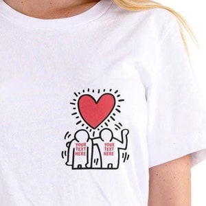 May include: White t-shirt with a black and white graphic of two figures holding a red heart. The text "YOUR TEXT HERE" is written on each figure.