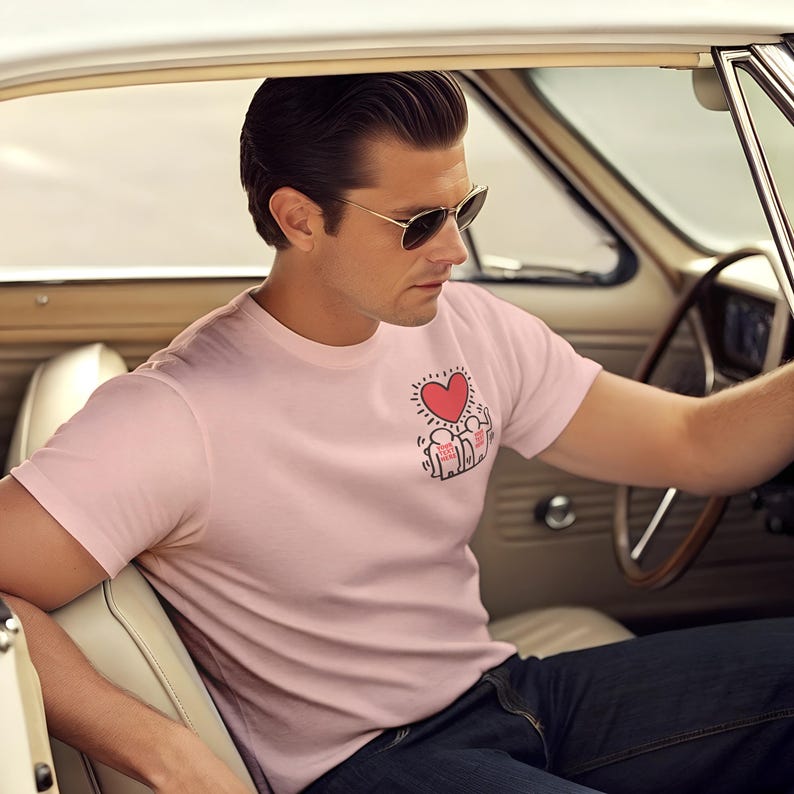 May include: A man wearing a pink t-shirt with a red heart and a graphic of people holding hands. He is wearing sunglasses and sitting in a car.