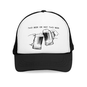 Könnte beinhalten: Schwarz-weiße Trucker-Cap mit dem Text "TWO BEER OR NOT TWO BEER". Die Grafik zeigt zwei Hände, die mit Bierkrügen anstoßen. Die Kappe hat einen schwarzen Schirm und eine Netzrückseite.