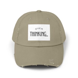 Può includere: Cappellino da baseball kaki consumato con visiera curva. Sul davanti, un rettangolo bianco con la scritta nera "OVER THINKING...". Il cappellino ha un aspetto vintage e usurato con bordi sfilacciati.