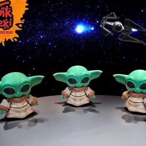 Może przedstawiać: Trzy zielone i brązowe figurki popularnej postaci fikcyjnej z franczyzy Star Wars. Figurki stoją na szarym tle na czarnym tle z białymi gwiazdami.