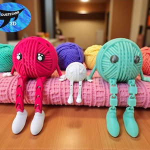 Könnte beinhalten: Eine Sammlung farbenfroher Garnkugelfiguren mit angebrachten Armen, Beinen und Füßen, die auf einer rosa Strickdecke liegen. Die Figuren sind in verschiedenen Farben gehalten, darunter Pink, Türkis, Lila, Gelb und Weiß. Die Figuren haben Augen und sind 3D-gedruckt.