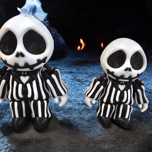 Könnte beinhalten: Zwei schwarz-weiß gestreifte Skelettfiguren mit schwarzen Augen und einer schwarzen Fliege. Die Figuren stehen auf einer dunkelblauen Oberfläche.