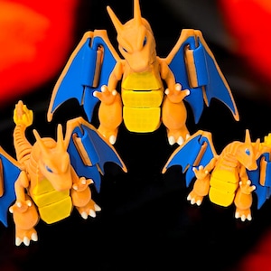 Könnte beinhalten: Drei orangefarbene und gelbe Charizard-Pokémon-Figuren mit blauen Flügeln. Die Figuren stehen in einer stehenden Position mit ausgebreiteten Flügeln.