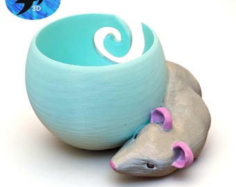 May include: A 3D-printed, light blue, spherical yarn bowl with a white swirl design. A gray mouse with pink ears and nose is attached to the bowl. The bowl has the text "duststarr 3D" on it.