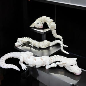 May include: Three white articulated dragon sculptures with detailed features, including scales and expressive faces. The dragons are posed on a black surface and a small black shelf, showcasing their flexible design and artistic craftsmanship.