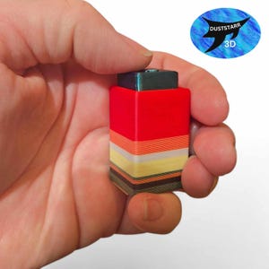 Op de afbeelding: Een klein, veelkleurig 3D-geprint object dat in een hand wordt gehouden. Het object is voornamelijk rood met gelaagde strepen van oranje, geel en bruin. Bovenop zit een donkergroene vierkant. De afbeelding bevat een logo "DUSTSTARR 3D".