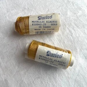 May include: Two spools of gold metallic elastic thread, each with a label that reads "Darice Metallic Elastic #10062-22 Gold 36 Yards Made in Japan". The spools are on a white, textured surface.