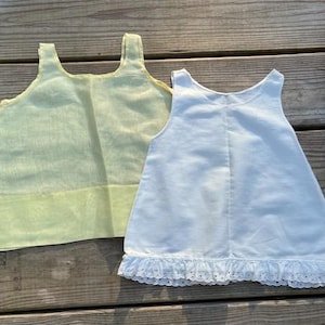 May include: Two sleeveless tops. One is a pale yellow-green, the other is white with a lace trim at the hem. Both have a simple, scoop neckline and are made of a lightweight fabric. The tops are laying on a weathered wooden surface.