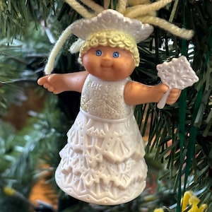 May include: A vintage Christmas ornament featuring a cherubic figure with blonde curls, blue eyes, and a white dress. The ornament is holding a white object and is suspended from a Christmas tree. The figure has a glittery bodice.