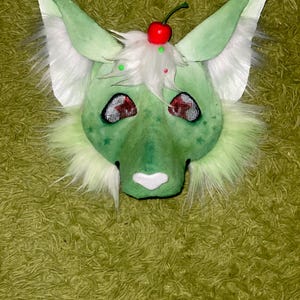 Puede incluir: Una m&aacute;scara peluda verde y blanca con orejas grandes, un hocico blanco y ojos rojos. La m&aacute;scara est&aacute; decorada con una cereza roja y pelusa blanca en la parte superior. La m&aacute;scara est&aacute; dise&ntilde;ada para parecerse a un animal de dibujos animados.