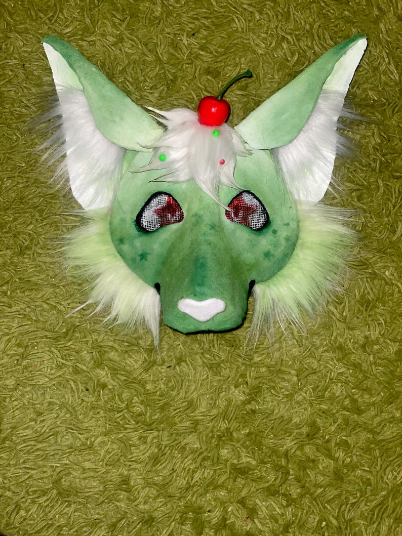 Puede incluir: Una m&aacute;scara peluda verde y blanca con orejas grandes, un hocico blanco y ojos rojos. La m&aacute;scara est&aacute; decorada con una cereza roja y pelusa blanca en la parte superior. La m&aacute;scara est&aacute; dise&ntilde;ada para parecerse a un animal de dibujos animados.