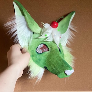 Puede incluir: Una m&aacute;scara de zorro peluda verde y blanca con grandes orejas puntiagudas, un hocico blanco y ojos de malla roja. La m&aacute;scara tiene una cereza encima y est&aacute; sujeta por una mano.